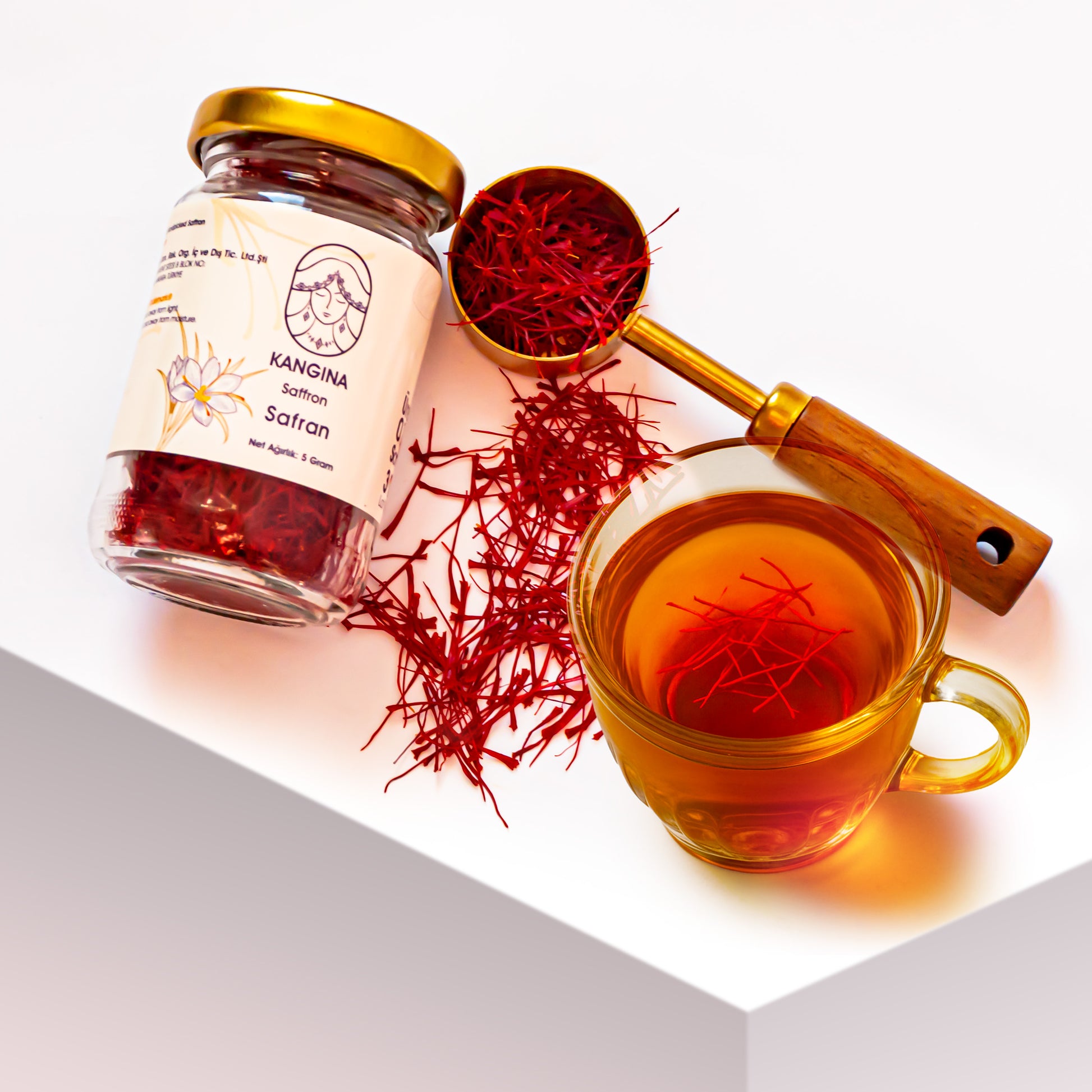 Kangina premium Afghan saffron threads top‑angle view of Kangina saffron jar, brass scoop filled with threads and a glowing cup of saffron tea on white backdrop